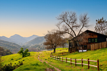 Serene southern California rural landscape with oak trees, fenced wooden hut on green meadow and mountains background.