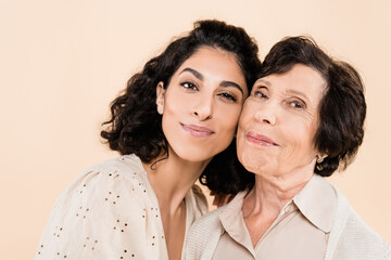 Smiling hipanic woman looking at camera near elderly mother isolated on beige, two generations of women