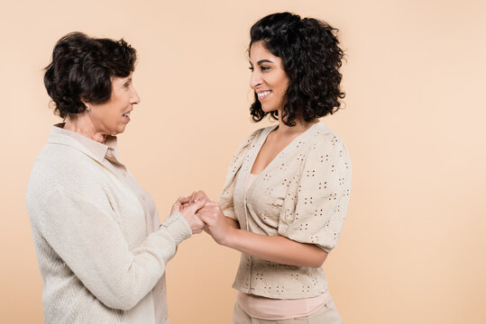 Smiling hispanic woman holding hands of elderly mother isolated on beige, two generations of women