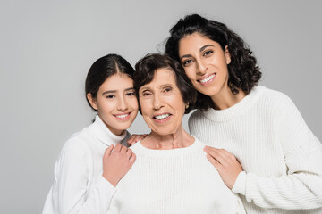 Smiling hispanic woman and daughter hugging elderly grandmother isolated on grey, three generations...