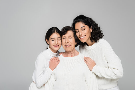 Smiling hispanic child and mother hugging senior grandmother with closed eyes isolated on grey, three generations of women