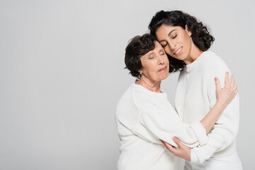 Senior woman embracing hispanic daughter with closed eyes isolated on grey, two generations of women