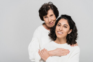 Hispanic woman smiling while touching hand of senior mother isolated on grey, two generations of women