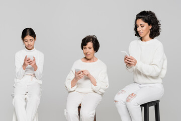 Hispanic women and kid using smartphones on chairs isolated on grey, three generations of women