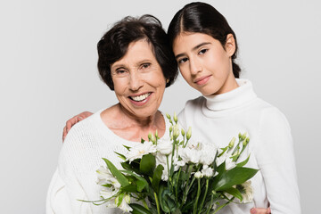 Smiling hispanic girl hugging granny with bouquet isolated on grey, two generations of women
