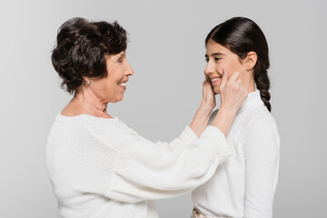 Smiling hispanic granny touching cheeks of granddaughter isolated on grey, two generations of women