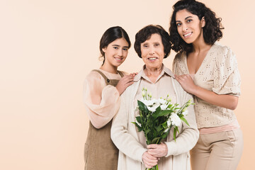 Hispanic mother and daughter hugging grandmother with flowers isolated on beige, three generations of women