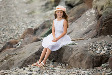 A cute smiling blonde girl 9 years old in a hat and white dress sits on the seashore.