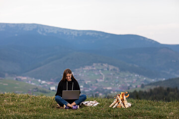 young female freelancer working on laptop in the mountains in the evening. Tourist girl sitting near campfire and having fun. Copy space.