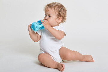 Little baby drinking water from blue bottle, looking aside while sitting on floor, posing barefoot...