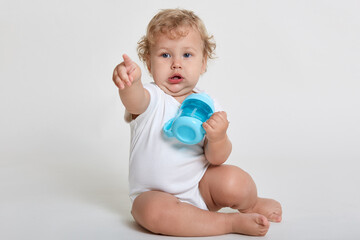 Cute baby wearing bodysuit holding feeding bottle in hands, pointing index finger away, having curious facial expression, posing isolated over light background.