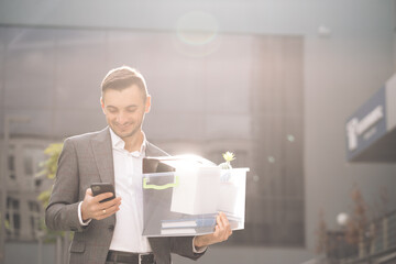Fototapeta na wymiar Happy businessman stand smiling use phone near business center. Businessman with box of personal stuff uses phone texting scrolling tapping. Portrait suit career male office handsome technology