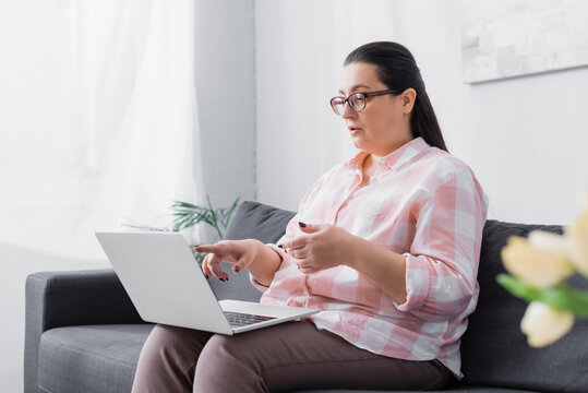 Plus size hispanic woman talking and gesturing while looking at laptop on couch at home on blurred foreground
