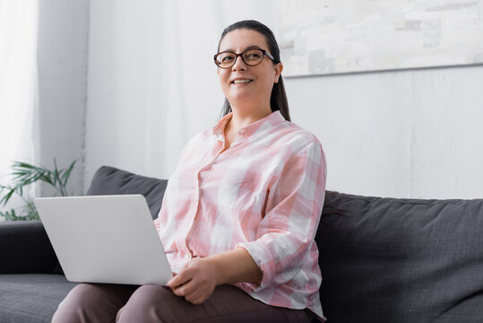 Smiling Plus Size Hispanic Woman With Laptop Looking Away While Sitting On Couch On Blurred Background