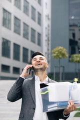 Happy fired man talking on phone with stuff in box. Speaking on telephone. Cellphone conversation. An employee between office buildings with a box and documents with a desk flower