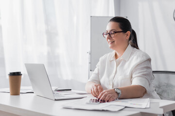 Happy plus size hispanic businesswoman looking at laptop during video call at workplace with papers on blurred foreground