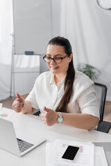 Smiling plus size hispanic businesswoman showing thumbs up while looking at laptop with blurred office on background