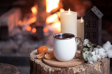Cozy composition with a cup, candles and tangerines on a blurred background of a burning fireplace.