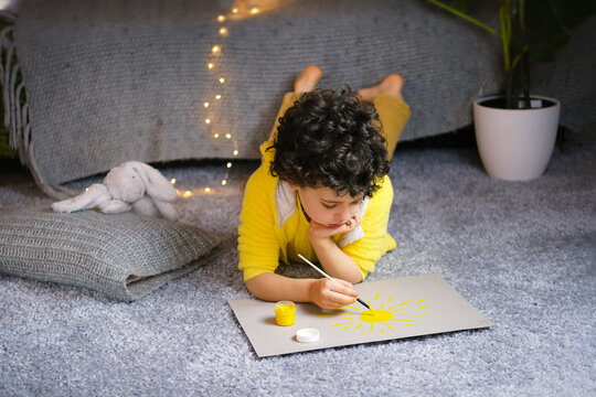 Little curly boy drawing yellow sun in gray interior.