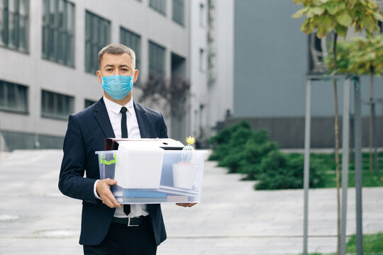 Young Sad Businessman In Protective Face Mask Walking Down The Stairs Outdoor With Box Of Stuff As Leaving Business Center. Unemployment Rate Growing Due Pandemic. Male Office Worker Lost Her Job