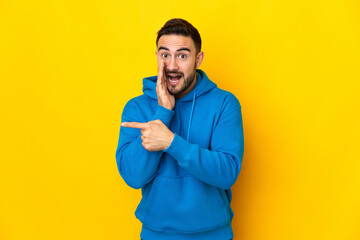Young caucasian handsome man isolated on yellow background pointing to the side to present a product and whispering something