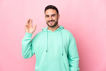 Young caucasian handsome man isolated on pink background saluting with hand with happy expression