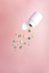 Many green pills and one orange are flying out of the bottle on a pink background.