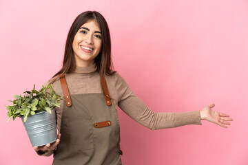 Young gardener girl holding a plant isolated on pink background extending hands to the side for inviting to come