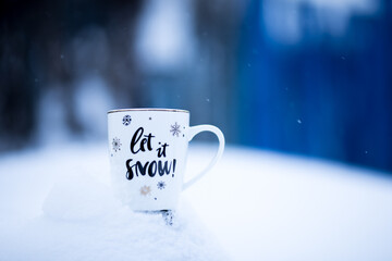 White Christmas cup on a snowdrift, blue background, outside. Mug with the inscription: "Let it snow"! Chrismas time, wintertime.