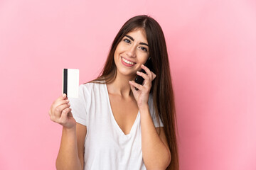 Young caucasian woman isolated on pink background keeping a conversation with the mobile phone and holding a credit card