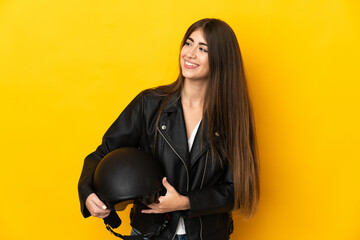 Young caucasian woman holding a motorcycle helmet isolated on yellow background looking to the side and smiling