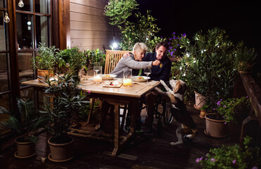 Senior couple in wheelchair having dinner in the evening on terrace, feeding dog.
