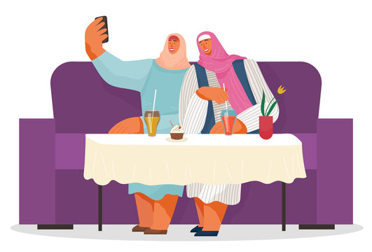 Two women friends wearing hijabs taking selfie together at phone. Arab women sitting at sofa in cafe or restaurant, drinking lemonade, eating cupcake. People spend leisure time. Isolated characters
