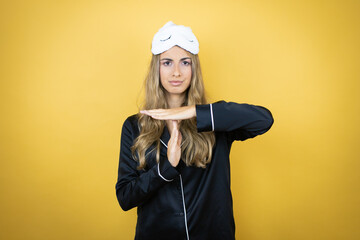 Young pretty woman wearing sleep mask and pajamas over isolated yellow background Doing time out gesture with hands, frustrated and serious face