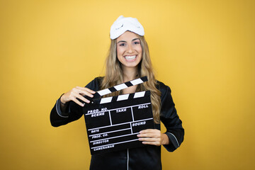 Young pretty woman wearing sleep mask and pajamas over isolated yellow background holding clapperboard very happy having fun