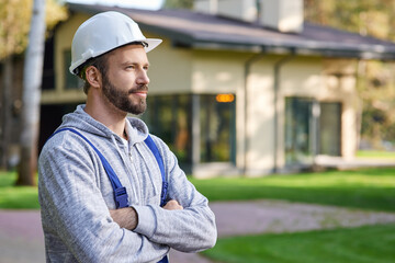 Close up of young male engineer in hard hat looking away, posing with arms crossed outdoors while...