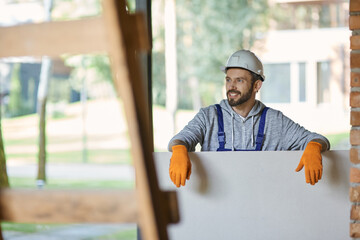 Handsome young male builder in hard hat looking positive, holding drywall while working at...