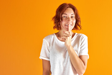 redhead woman has a secret, show keep silence gesture, smile looking at camera isolated over orange...