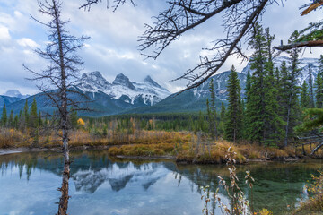 Fototapeta na wymiar Snow capped The Three Sisters trio peaks mountain with blue sky and white clouds reflect on water surface in autumn. Beautiful natural scenery landscape at Canmore, Canadian Rockies, Alberta, Canada.