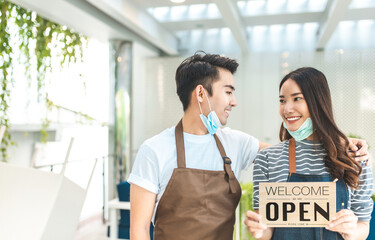 Business owner attractive young Asian couple in apron hanging we're open sign on  welcoming clients to new cafe. Happy waiter with protective face mask holding open sign while stand at cafe .