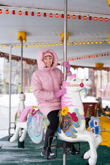 A young woman in a pink down jacket rides in winter in a city park on a children's carousel