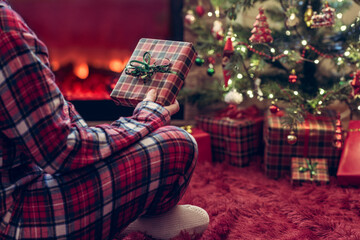 Fototapeta na wymiar Woman in pajamas with christmas gift box in hand sitting on fluffy plaid near fireplace and christmas tree.