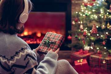 Woman in headphones with christmas gift box in hand sitting on fluffy plaid near fireplace and...