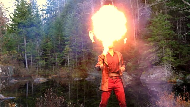 Portrait of handsome young man near lake in forest wearing metal horn holding illuminated fire stick spitting fire from mouth during fire jamming 