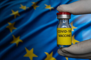 Bottle vial vaccine on flag European Union background. COVID-19 Pandemic Coronavirus concept. Hand is holding covid vaccine bottle. Concept of Covid vaccination campaign in Europe.