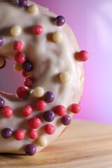 White icing donut with small colored pieces of chocolate on a wooden table against purple background