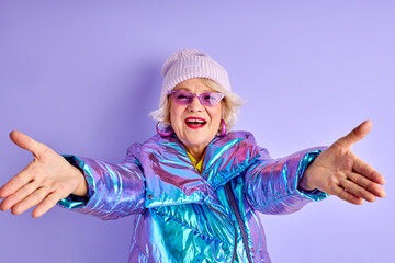 cheerful elderly woman with spread arms look at camera, going to hug, wearing winter coat hat and sunglasses