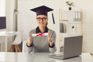 Happy online courses graduate sitting at desk with laptop, holding her diploma and smiling