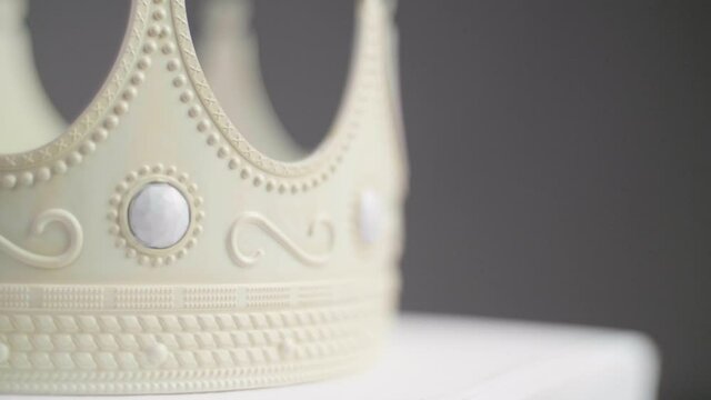 Close up of a beige crown on a pedestal with ornaments and white pearl jewels with peaks jagged out of plastic. With a neutral grey background. Smooth backwards dolly camera movement. Shot in 4K.	