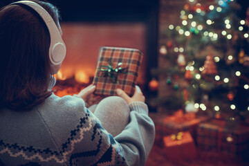 Woman in headphones with christmas gift box in hand sitting on fluffy plaid near fireplace and...